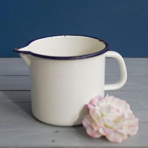 Könnte beinhalten: Weißer Emaille-Krug mit blauem Rand und Henkel. Der Krug steht auf einer hellgrauen Holzoberfläche mit einer rosa Blume.