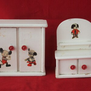 May include: A white wooden dollhouse furniture set with two pieces. The larger piece is a two-door cabinet with red knobs and cartoon animal illustrations. The smaller piece is a vanity with a shelf, two drawers with red knobs, and a cartoon animal illustration.