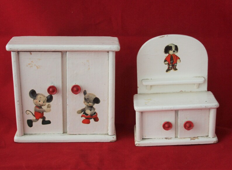 May include: A white wooden dollhouse furniture set with two pieces. The larger piece is a two-door cabinet with red knobs and cartoon animal illustrations. The smaller piece is a vanity with a shelf, two drawers with red knobs, and a cartoon animal illustration.