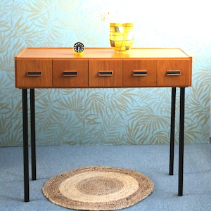 Op de afbeelding: Een houten console in het midden van de eeuw met vier lades en zwarte metalen poten. Een gele en zwarte vaas en een kleine klok staan bovenop. Een ronde, geweven vloerkleed ligt op de vloer.