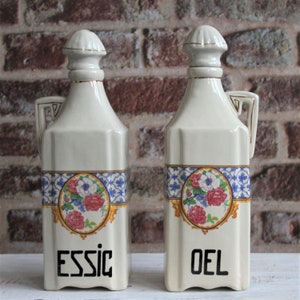 May include: Two white ceramic bottles with floral designs and gold accents. The bottles are labeled "ESSIG" and "OEL".
