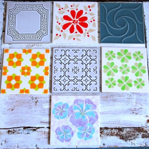 Könnte beinhalten: Eine Sammlung von neun quadratischen Keramikfliesen mit verschiedenen Designs. Die Fliesen zeigen florale Muster, geometrische Formen und ein Wirbeldesign in Rot-, Orange-, Grün-, Blau- und Schwarztönen. Die Fliesen sind auf einer weißen, verwitterten Holzoberfläche angeordnet.
