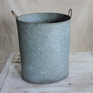 Könnte beinhalten: Ein galvanisierter Metalltopf mit abgerundeter ovaler Form und zwei Drahtgriffen. Der Topf ist aus strukturiertem Metall und hat eine hellgraue Farbe.