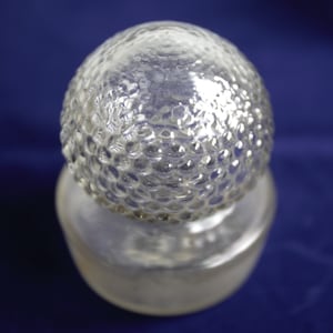 Op de afbeelding: Een heldere glazen fles met een getextureerde, bolvormige stopper. De stopper heeft een belachtig patroon. De fles staat op een blauw oppervlak. De fles is waarschijnlijk voor decoratieve doeleinden.