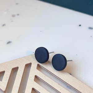 Può includere: Due orecchini a bottone neri opachi con perni d'argento. Gli orecchini sono circolari e poggiano su una superficie di legno chiaro con intagli geometrici. Lo sfondo è bianco e verde acqua scuro.
