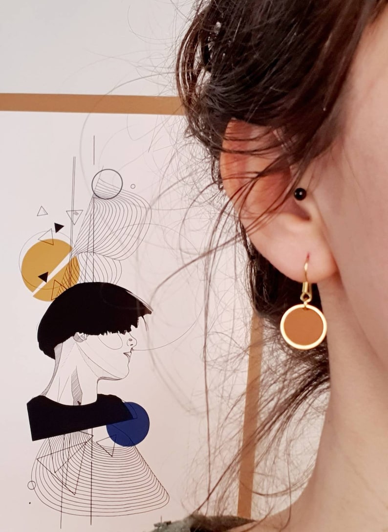 Puede incluir: Un par de pendientes de aro dorados con un colgante de disco marr&oacute;n. Los pendientes cuelgan de la oreja de una persona.