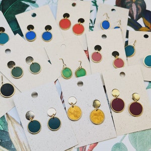 Puede incluir: Surtido de pendientes redondos de colores con detalles dorados. Los pendientes se muestran sobre una superficie blanca con un fondo de hojas verdes.