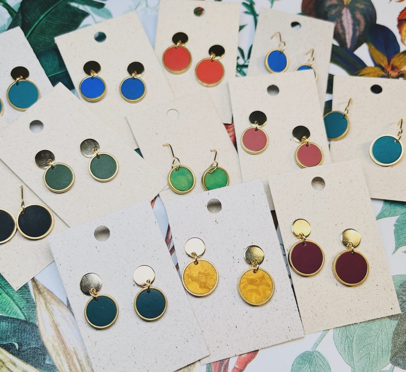 Puede incluir: Surtido de pendientes redondos de colores con detalles dorados. Los pendientes se muestran sobre una superficie blanca con un fondo de hojas verdes.