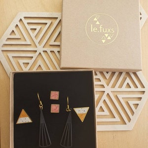 Puede incluir: Un conjunto de pendientes en una caja negra. Los pendientes est&aacute;n hechos de metal negro con un dise&ntilde;o geom&eacute;trico. Los pendientes se emparejan con peque&ntilde;os pendientes cuadrados con un dise&ntilde;o moteado rosa y blanco. La caja est&aacute; etiquetada con "le.fuxs".