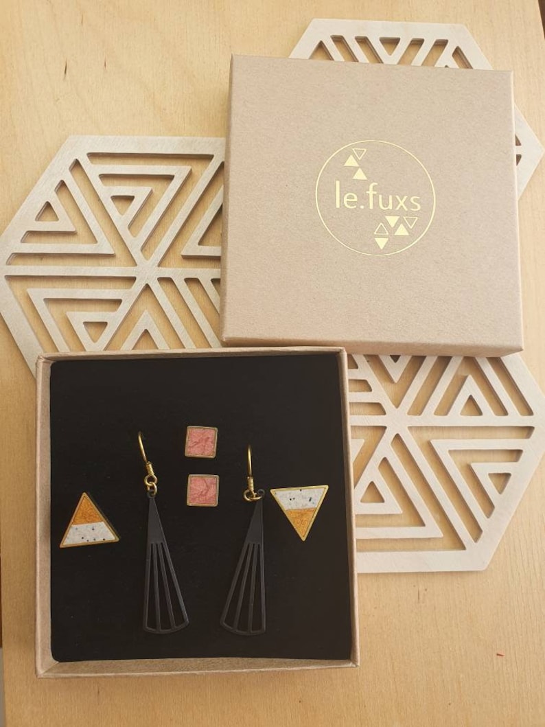 Puede incluir: Un conjunto de pendientes en una caja negra. Los pendientes est&aacute;n hechos de metal negro con un dise&ntilde;o geom&eacute;trico. Los pendientes se emparejan con peque&ntilde;os pendientes cuadrados con un dise&ntilde;o moteado rosa y blanco. La caja est&aacute; etiquetada con "le.fuxs".