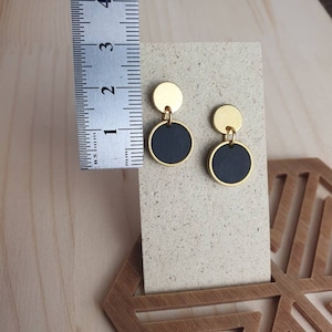Puede incluir: Un par de pendientes de oro y negro con un dise&ntilde;o simple y moderno. Cada pendiente tiene un peque&ntilde;o c&iacute;rculo dorado coronado por un c&iacute;rculo negro m&aacute;s grande.