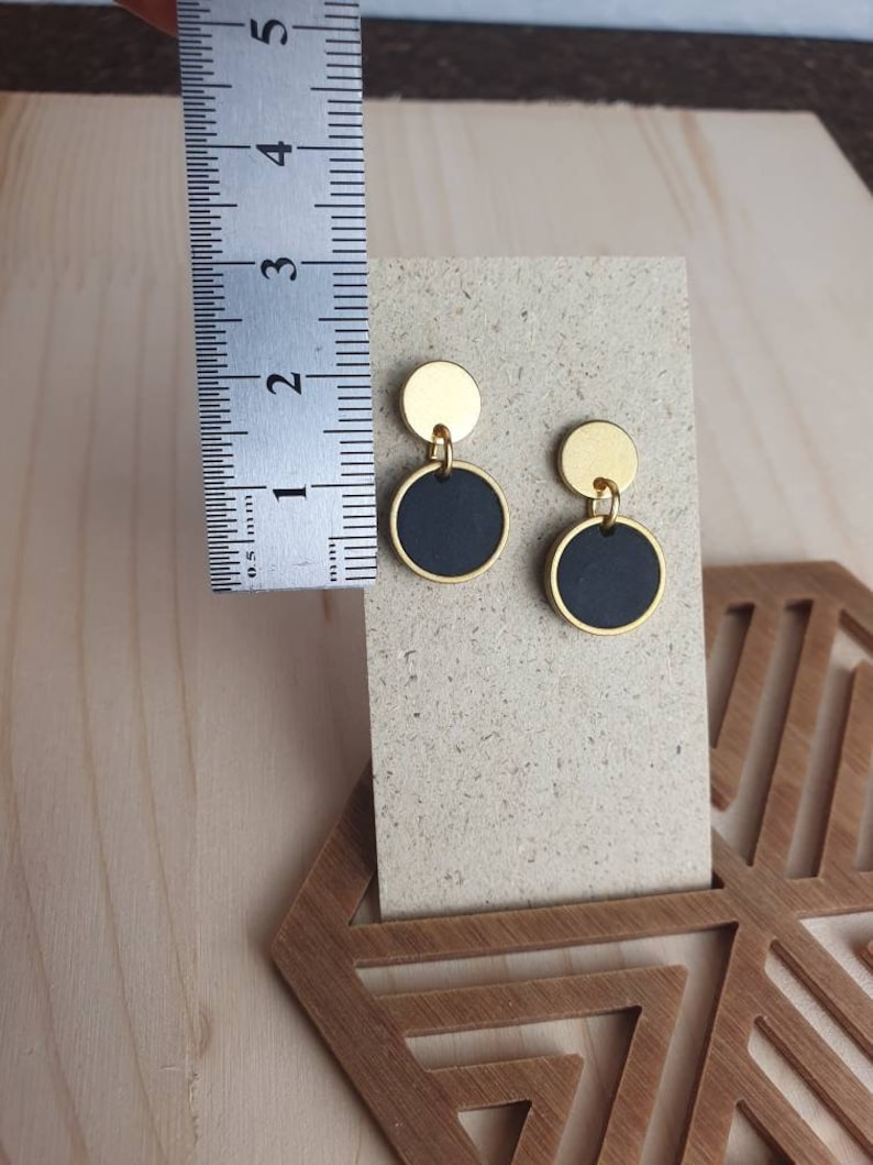 Puede incluir: Un par de pendientes de oro y negro con un dise&ntilde;o simple y moderno. Cada pendiente tiene un peque&ntilde;o c&iacute;rculo dorado coronado por un c&iacute;rculo negro m&aacute;s grande.
