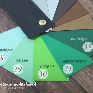 May include: A color chart of green cardstock samples. The colors are pastel green, pistachio, grass green, Christmas green, and dark green. Each color is labeled with a number and a German name. The chart is on a wooden surface.
