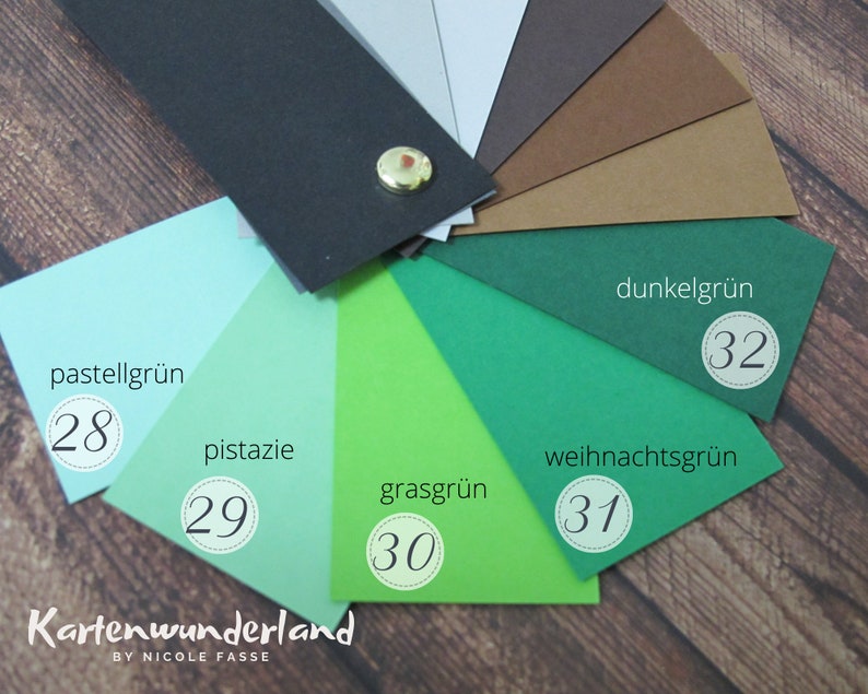 May include: A color chart of green cardstock samples. The colors are pastel green, pistachio, grass green, Christmas green, and dark green. Each color is labeled with a number and a German name. The chart is on a wooden surface.
