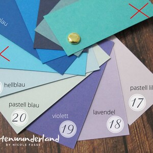 May include: A color chart with 10 different shades of blue, purple, and lavender. The colors are labeled with German words and numbers. The chart is on a wooden surface.