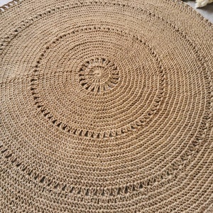 Puede incluir: Una alfombra redonda de yute en tonos naturales, con un dise&ntilde;o en espiral. La alfombra presenta una serie de c&iacute;rculos conc&eacute;ntricos, con un centro detallado y un borde festoneado. La textura tejida es claramente visible.