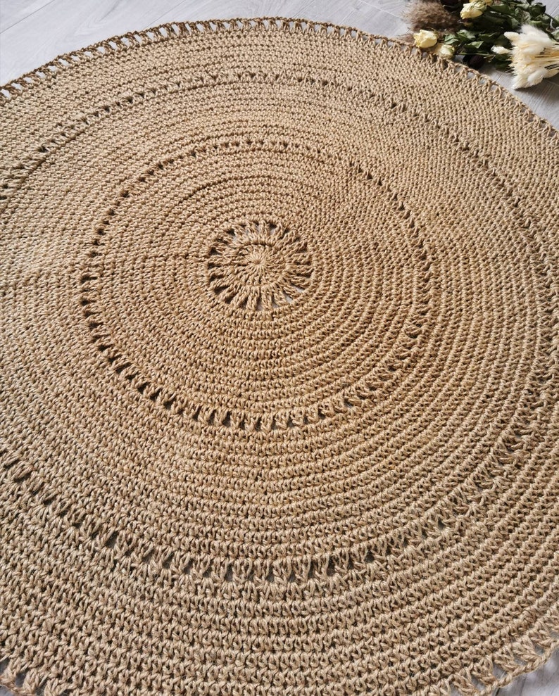 Puede incluir: Una alfombra redonda de yute en tonos naturales, con un dise&ntilde;o en espiral. La alfombra presenta una serie de c&iacute;rculos conc&eacute;ntricos, con un centro detallado y un borde festoneado. La textura tejida es claramente visible.