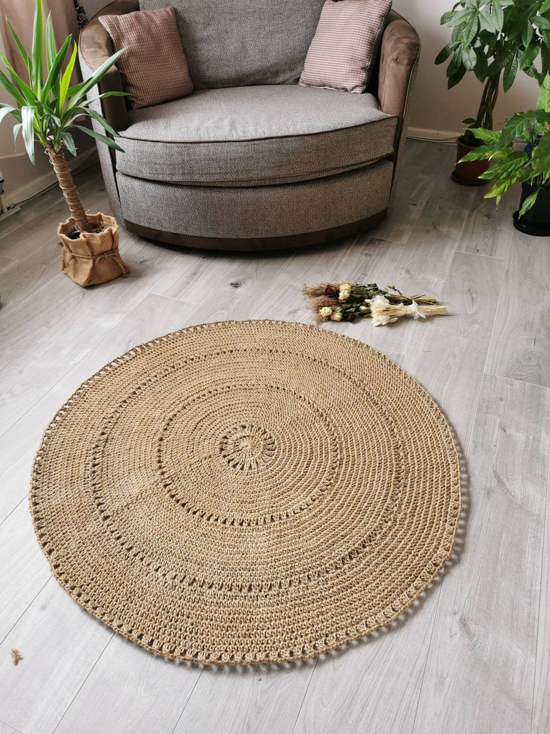 Puede incluir: Una alfombra redonda de ganchillo beige con un dise&ntilde;o en espiral. La alfombra est&aacute; hecha de una fibra natural y tiene una superficie texturizada. La alfombra est&aacute; colocada sobre un suelo de madera gris claro. La alfombra tiene unos 122 cm de di&aacute;metro.