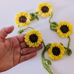 May include: A crocheted sunflower garland with yellow petals, brown centres, and green leaves. This handmade garland is a decorative item, ideal for adding a touch of nature. The sunflowers are approximately 7.6 cm in diameter.