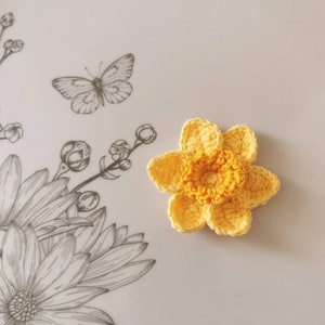 May include: A crocheted yellow daffodil flower with a yellow center. The flower is on a white background with a black and white floral design.