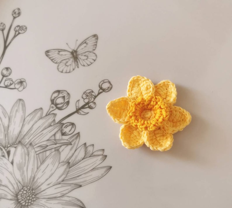 May include: A crocheted yellow daffodil flower with a yellow center. The flower is on a white background with a black and white floral design.