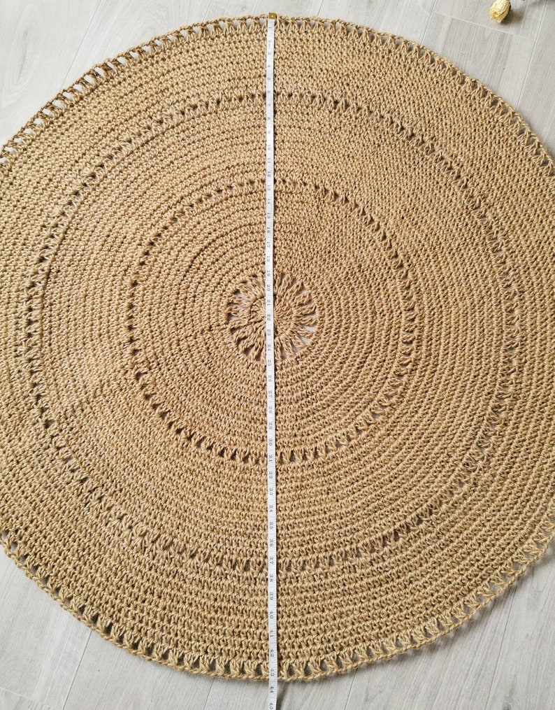 Puede incluir: Una alfombra redonda de yute en tono natural, con un di&aacute;metro aproximado de 120 cm. La alfombra presenta c&iacute;rculos conc&eacute;ntricos y patrones decorativos calados. Una cinta m&eacute;trica se coloca en el centro. La alfombra est&aacute; sobre una superficie clara.