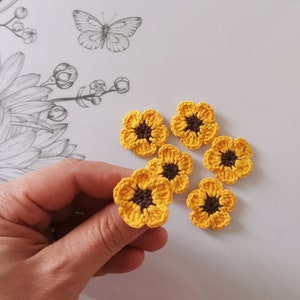 May include: Six crocheted sunflower embellishments in yellow and brown. The flowers are small and delicate, perfect for adding a touch of whimsy to crafts.