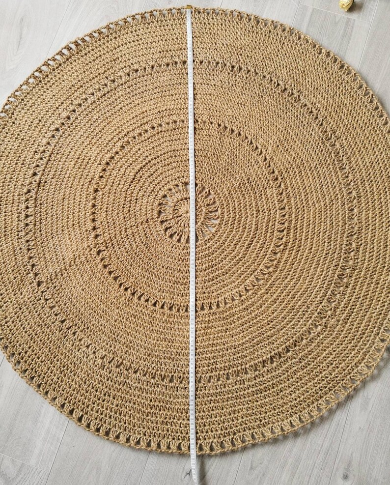 Puede incluir: Una alfombra redonda de yute en tono natural, con dise&ntilde;o de c&iacute;rculos conc&eacute;ntricos. Una cinta m&eacute;trica indica un di&aacute;metro de aproximadamente 152 cm. La alfombra tiene una superficie texturizada y un borde decorativo.