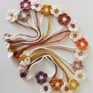 May include: A collection of crocheted flower embellishments made with white, pink, orange, and brown yarn. The flowers are attached to long lengths of yarn.