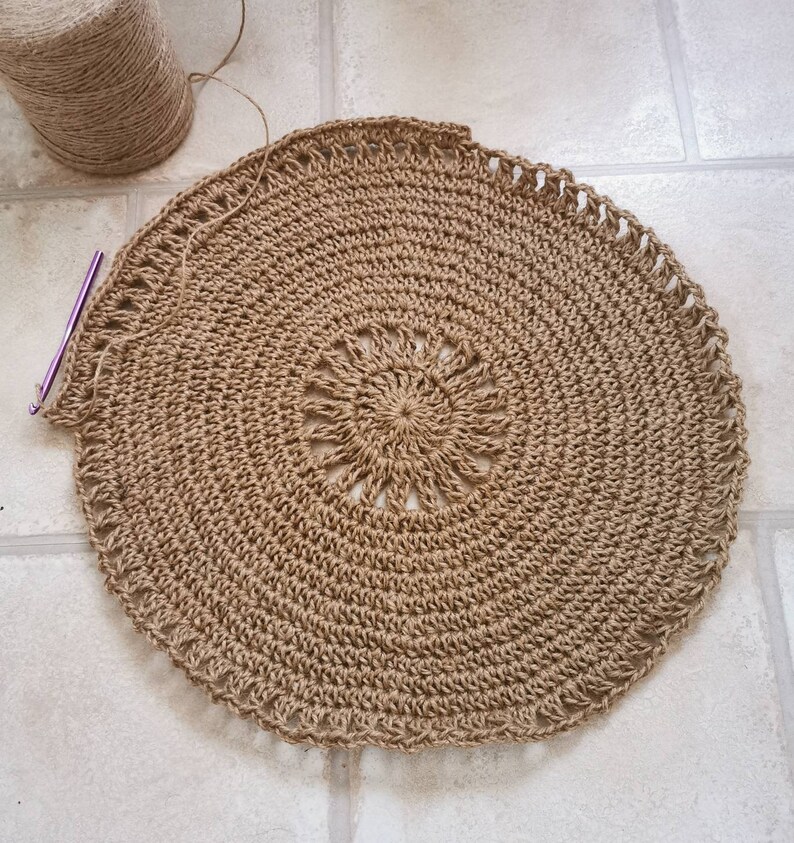 Puede incluir: Proyecto de ganchillo circular inacabado en tonos marr&oacute;n y beige. El centro presenta un dise&ntilde;o de sol. Se ven un ganchillo y un ovillo de hilo.