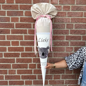Peut inclure: Un cornet scolaire blanc et beige avec le nom "Lieke" et le texte "Schulkind 2025". Le cornet est décoré d'un koala et de rubans roses et beiges. Le cornet est tenu devant un mur de briques.