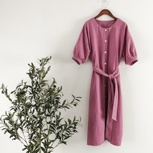 May include: A dusty rose linen dress with a button-up front and a self-tie belt. The dress has a relaxed fit and a slightly puffed sleeve.