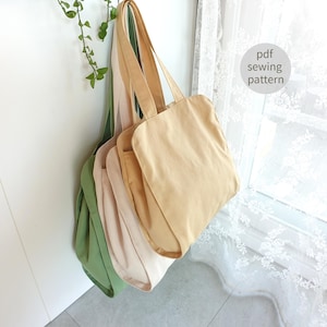Könnte beinhalten: Eine Sammlung von Canvas-Tragetaschen in verschiedenen neutralen Farben, darunter Beige, Hellrosa und Grün, die an einer weißen Wand hängen. Die Taschen haben lange Henkel und ein schlichtes, rechteckiges Design. Der Text auf dem Bild lautet "pdf sewing pattern".