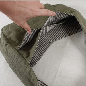 May include: An olive green fabric bag featuring an open top design. The interior is lined with a contrasting white and dark gray striped fabric, revealing two internal pockets visible within the main compartment of the bag.