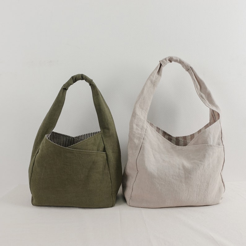 May include: Two hobo-style shoulder bags made of fabric. One bag is olive green with a dark striped interior lining, and the other is beige with a light brown striped interior lining. Both bags feature a single knotted handle and an exterior pocket.