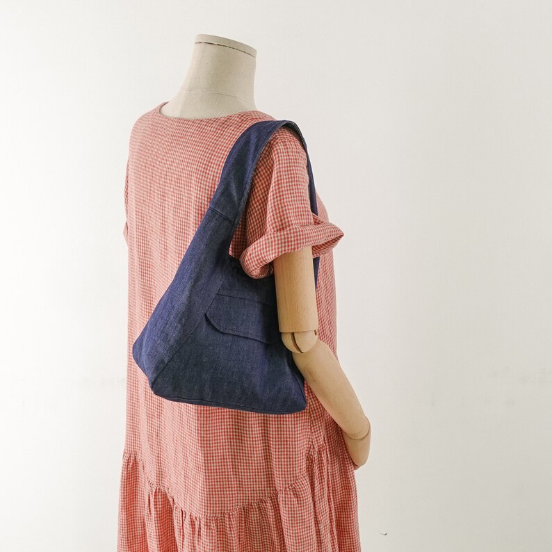 May include: A blue denim tote bag with a small pocket on the front. The bag is being worn by a mannequin wearing a red and white gingham dress.