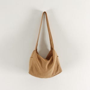 May include: A tan hobo-style shoulder bag crafted from a soft, textured fabric. The bag features a single wide shoulder strap, a slouchy, unstructured main compartment, and visible side pockets, presenting a simple and functional design.