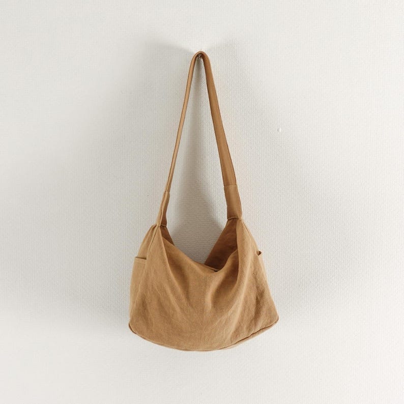 May include: A tan hobo-style shoulder bag crafted from a soft, textured fabric. The bag features a single wide shoulder strap, a slouchy, unstructured main compartment, and visible side pockets, presenting a simple and functional design.