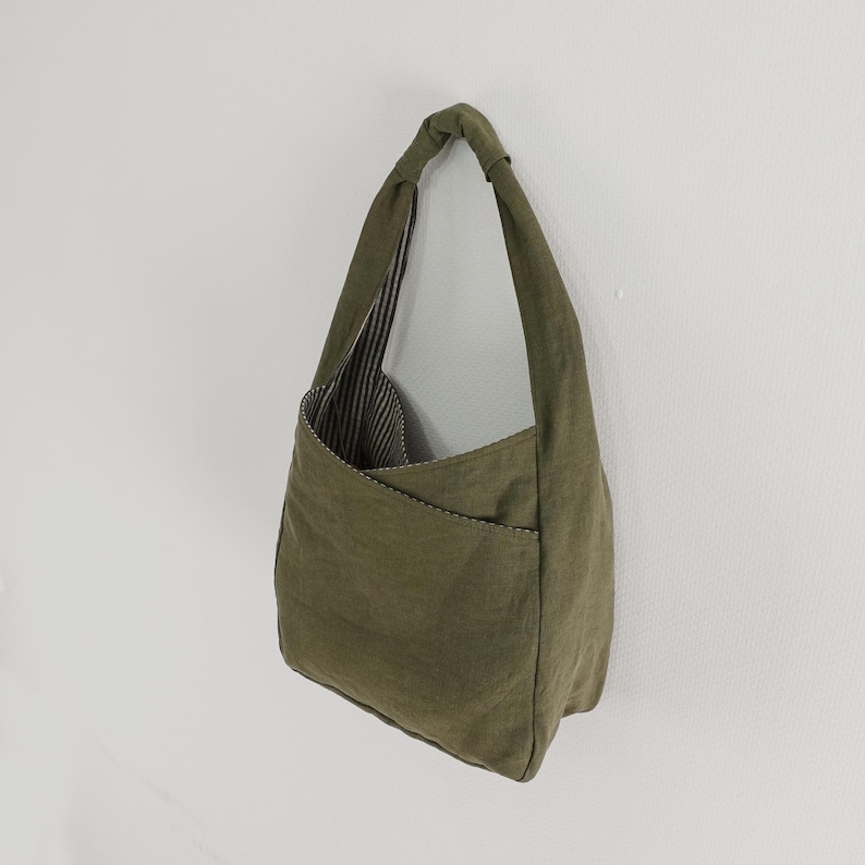 May include: An olive green hobo bag featuring a single integrated shoulder strap and a diagonal exterior pocket. The interior lining is visible and displays a black and white vertical striped pattern, contrasting with the solid green exterior fabric.