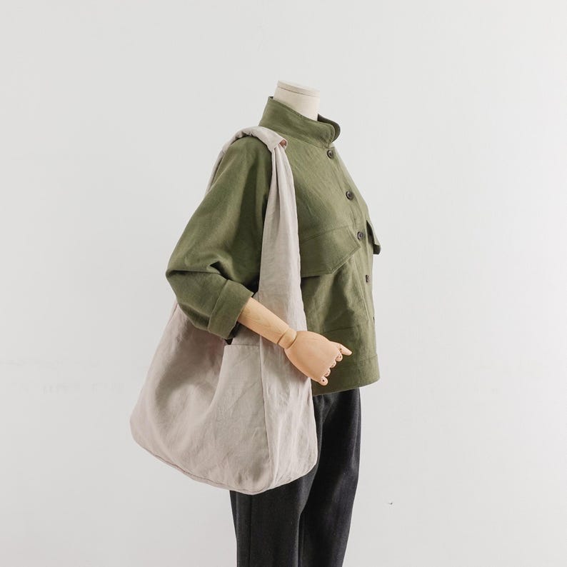 May include: A large, light beige hobo-style shoulder bag with a wide strap, worn over the shoulder. The bag is made of a solid-colored fabric and features a slouchy, unstructured shape. It is paired with an olive green button-up jacket and dark pants.