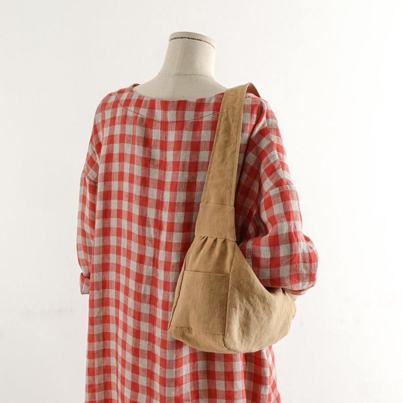 May include: A long-sleeved dress featuring a red and white gingham check pattern, worn with a tan hobo-style shoulder bag. The bag has a wide strap, a gathered top section, and a small exterior pocket on the front, resting against the fabric.