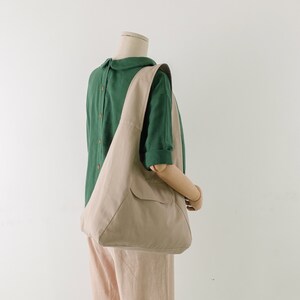 May include: A beige canvas tote bag with a single pocket on the front. The bag is being worn by a mannequin wearing a green collared shirt.