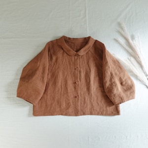 May include: A brown linen shirt with a peter pan collar and button closure. The shirt has a boxy fit and three-quarter sleeves.