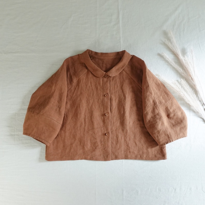 May include: A brown linen shirt with a peter pan collar and button closure. The shirt has a boxy fit and three-quarter sleeves.