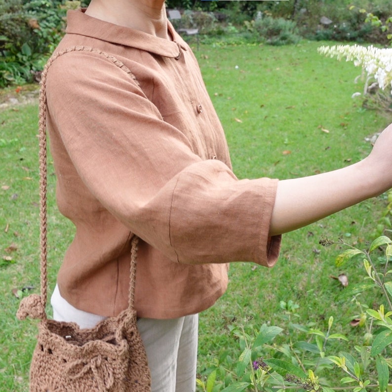May include: A brown linen shirt with a collared neckline and three-quarter sleeves. The shirt is worn by a person with a crocheted bag slung over their shoulder.