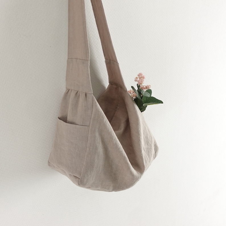 May include: A beige linen tote bag with a single pocket on the side. The bag is hanging from a white wall and has a small bouquet of pink flowers tucked inside.