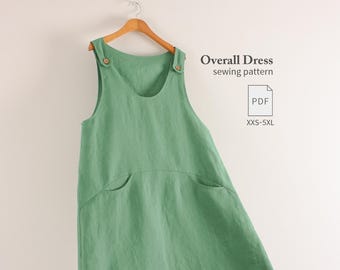 May include: A sage green overall dress hanging on a wooden hanger. The dress features two shoulder straps with small brown buttons and two front pockets. Text on the right reads Overall Dress sewing pattern, PDF, and XXS-5XL.