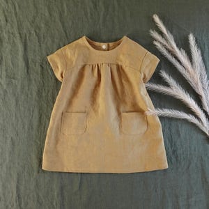 May include: A short-sleeved, tan-coloured dress featuring a round neckline with a single white button closure at the back, a gathered bodice, and two square patch pockets on the front. The garment has a simple, A-line silhouette.