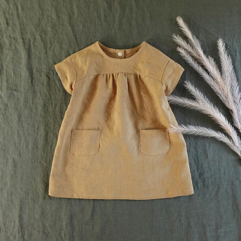 May include: A short-sleeved, tan-coloured dress featuring a round neckline with a single white button closure at the back, a gathered bodice, and two square patch pockets on the front. The garment has a simple, A-line silhouette.
