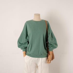 Könnte beinhalten: Ein salbeigrünes Langarm-Sweatshirt mit Rundhalsausschnitt und voluminösen Puffärmeln mit eng anliegenden Bündchen. Das Kleidungsstück wird mit einer weißen Hose und einer geflochtenen braunen Umhängetasche an einer Schaufensterpuppe präsentiert.